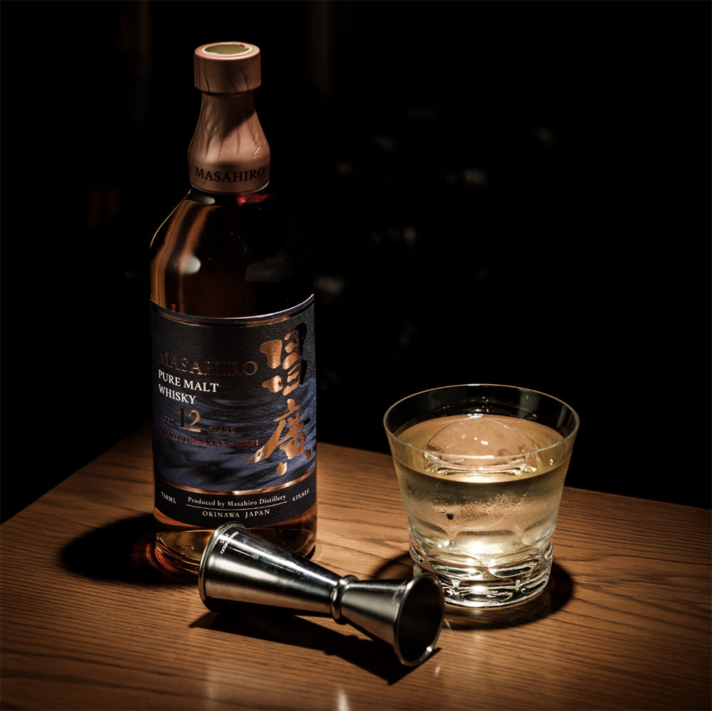 A bottle of Masahiro Pure Malt 12 Year Old Sherry Cask Japanese Whisky sits on a wooden table beside a glass of whisky with ice and a metal jigger, all highlighted by focused lighting.