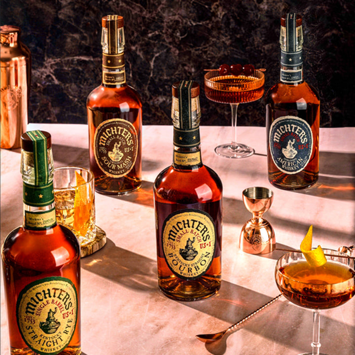 Michter's US*1 Kentucky Straight Bourbon Whiskey bottle is displayed on a marble surface with cocktails in elegant glasses, a copper jigger, and a mixing glass, set against a dark marble background.