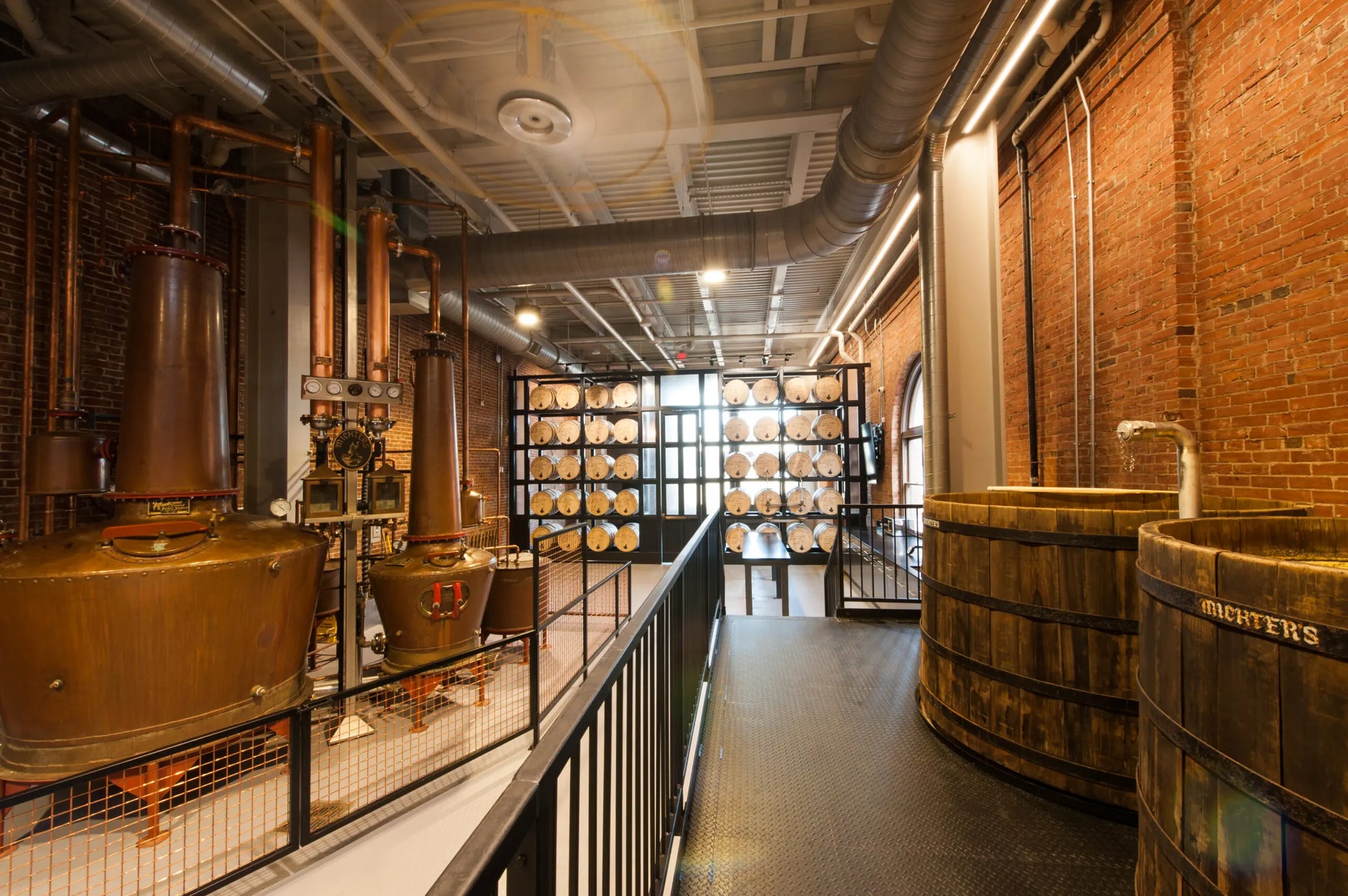 Industrial interior with large copper stills and wooden barrels in a distillery setting.