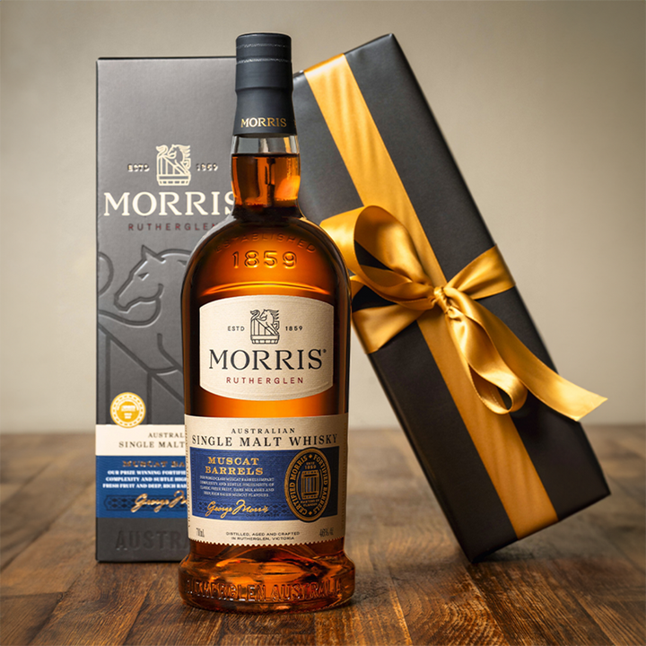 A bottle of Morris Whisky Muscat Barrel Australian Single Malt Whisky stands before its box beside a black gift box with a gold ribbon, all elegantly arranged on a wooden surface.