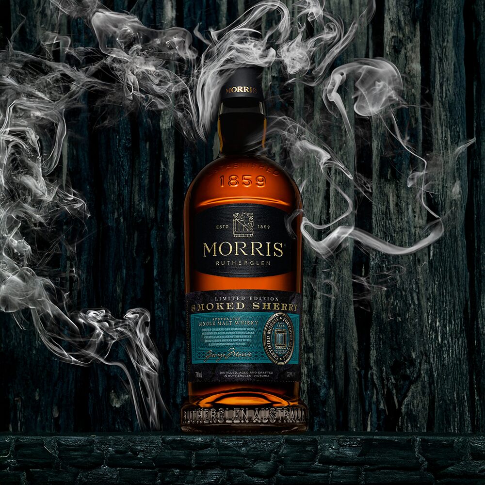 A bottle of Morris Whisky Smoked Sherry Australian Single Malt Whisky stands upright against charred wood, swirling smoke hinting at its rich, smoky character from finishing in smoked sherry casks.