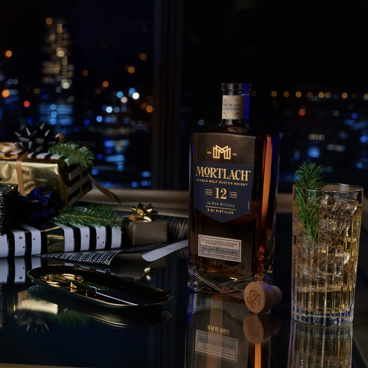 A bottle of Mortlach 12 Year Old Single Malt Scotch Whisky rests on a table with a glass, cork, wrapped gifts, and pine sprigs, set before a night city skyline—evoking the spirit of Speyside, Scotland.