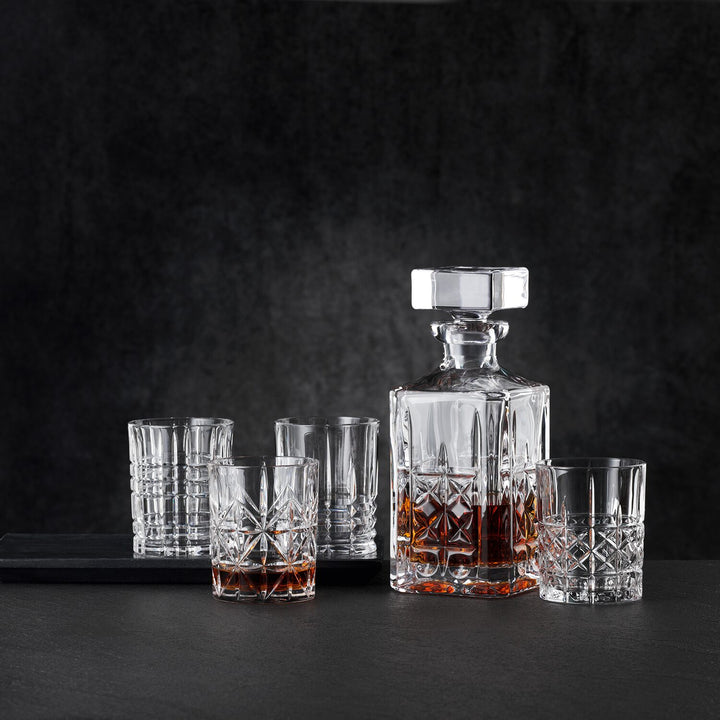The Nachtmann Highland Decanter Set features a crystal whisky decanter filled with amber liquor, accompanied by six matching empty glasses, all arranged against a dark, textured background.