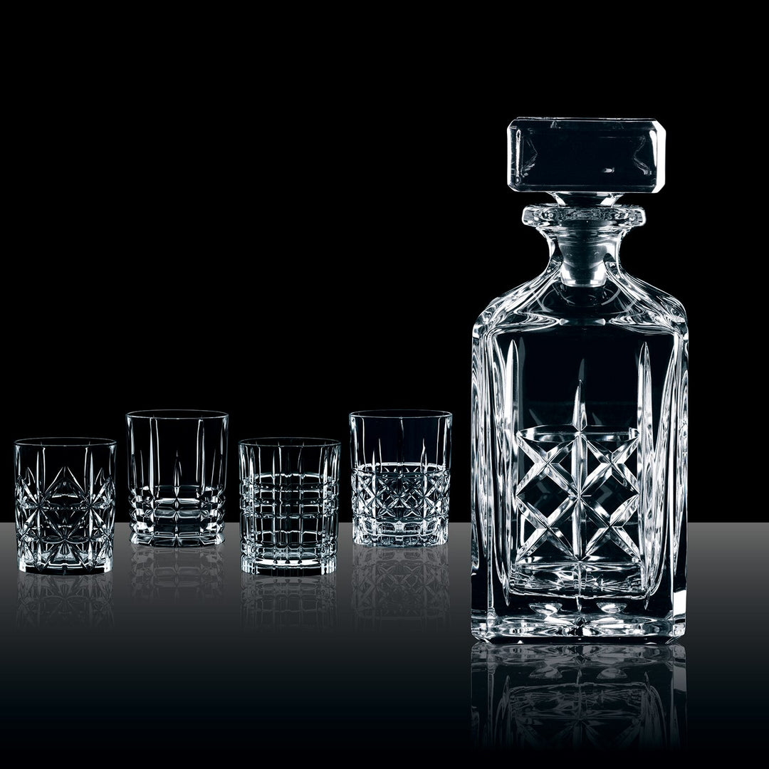 The Nachtmann Highland Decanter Set features a crystal whisky decanter with a square stopper and four patterned glasses, arranged on a reflective surface against a black background.