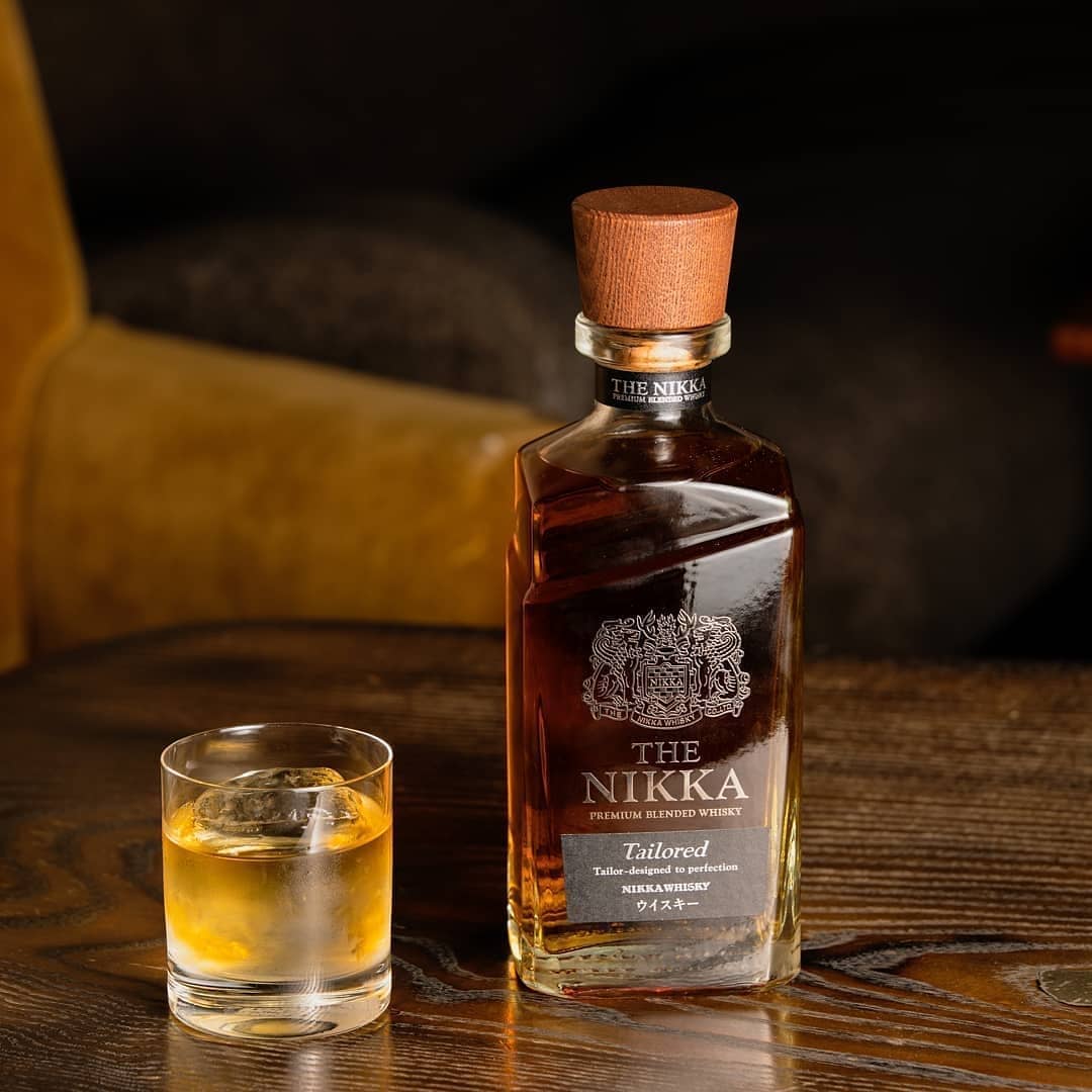 A bottle of Nikka Tailored Blended Japanese Whisky with a wooden cap stands on a wooden table beside a whisky glass with ice, while a brown chair is visible in the background.