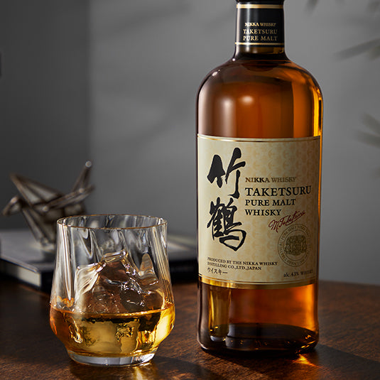 A bottle of Nikka Taketsuru Pure Malt Japanese Whisky sits beside a glass of whisky with ice on a wooden table, while a blurred object and a closed book in the background create an inviting atmosphere.
