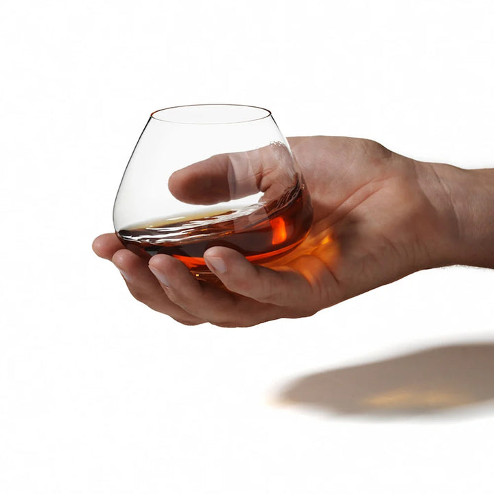 A hand holds a Normann Copenhagen Cognac Glass, partially filled with amber liquid, showcasing the modern design of the 2-piece glass set against a plain white background.