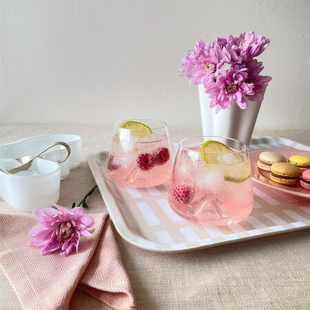 Two pink drinks are served in Normann Copenhagen Whisky Glasses (set of 2) with raspberries and lime, arranged on a tray beside colorful macarons, a white vase with pink flowers, and a folded pink napkin topped with a flower.