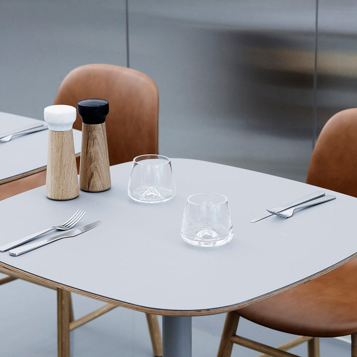 A modern dining table with two tan chairs, set with Normann Copenhagen Whisky Glasses (2 pcs), a fork and knife on either side, and two wooden salt and pepper grinders with white and black tops.