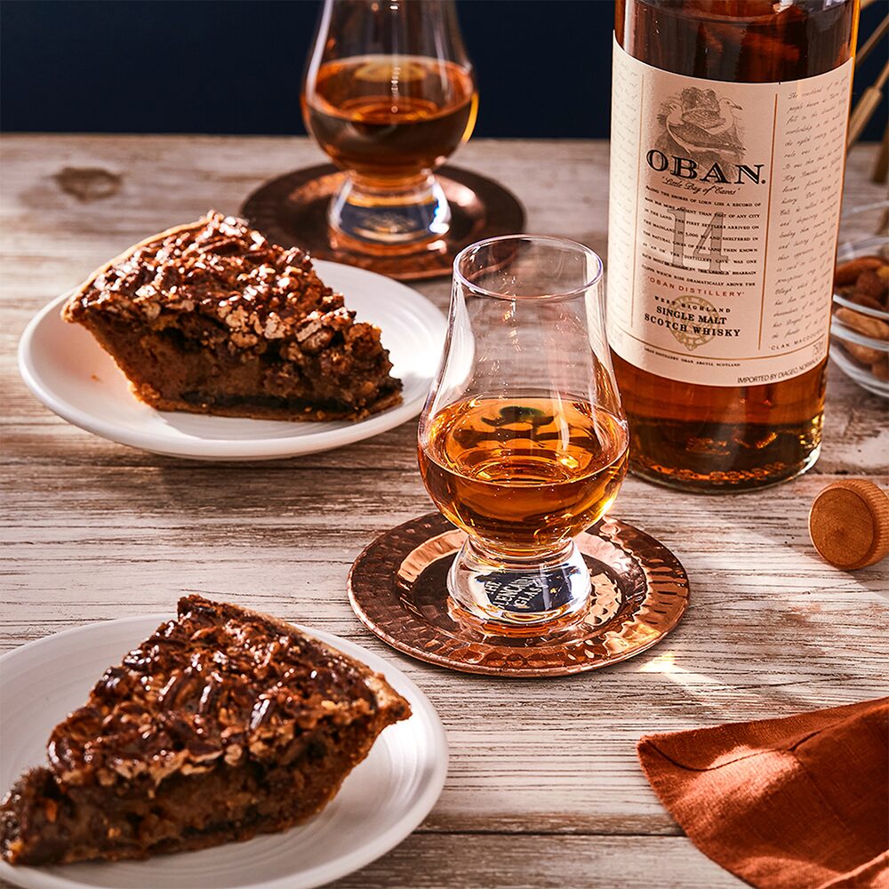 Two slices of pecan pie and glasses of Oban 14 Year Old Single Malt Scotch Whisky sit on white plates at a rustic wooden table, accompanied by a copper coaster and the whisky bottle’s cork.