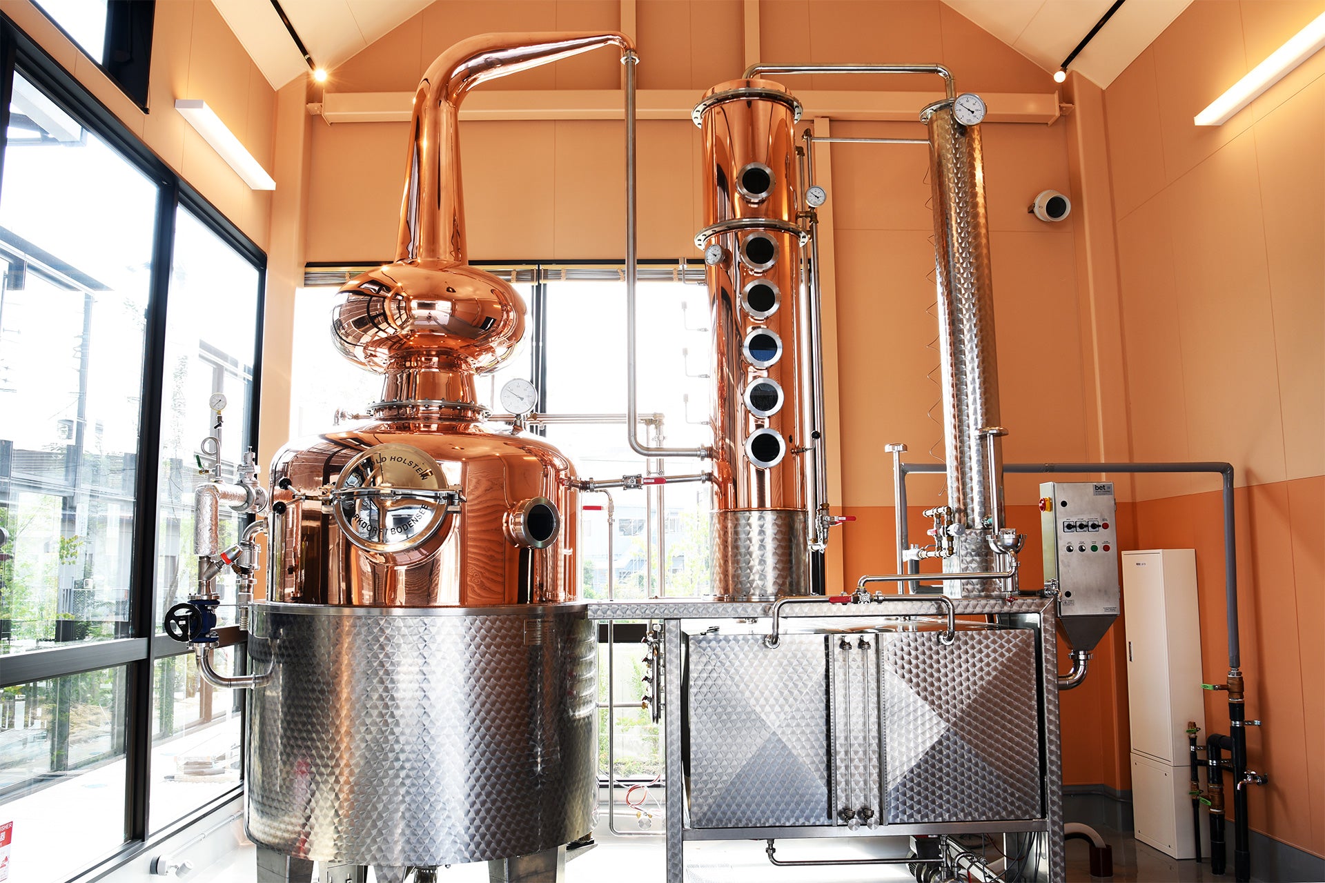 Copper stills in a distillery with large windows.