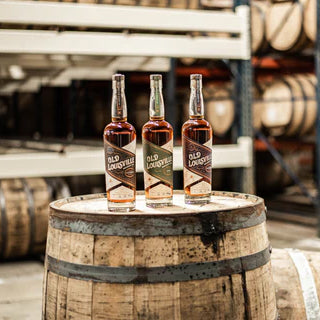 Three bottles of Old Louisville Whiskey Co. Straight Bourbon Whiskey Batch 2 rest on a wooden barrel, with blurred barrels and shelves in the background, evoking the authentic feel of a small-batch bourbon distillery.