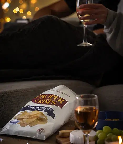 A bag of Proper Crisps Marlborough Sea Salt, cheese, grapes, and wine are arranged on a table with blurred lights. Someone relaxes on a sofa, savoring wine and enjoying the moment.