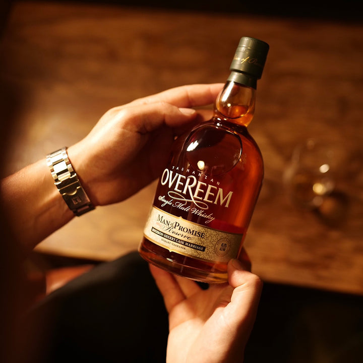 A person holds a bottle of Overeem Man of Promise 2023 Australian Single Malt Whisky with both hands over a wooden table, while a glass of whisky sits blurred in the background and the person wears a silver watch.