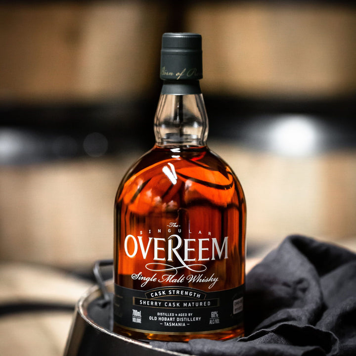 A bottle of Overeem Sherry Cask Matured – Cask Strength Australian Single Malt Whisky rests on a reflective surface beside a dark cloth, with blurred wooden barrels visible in the background.