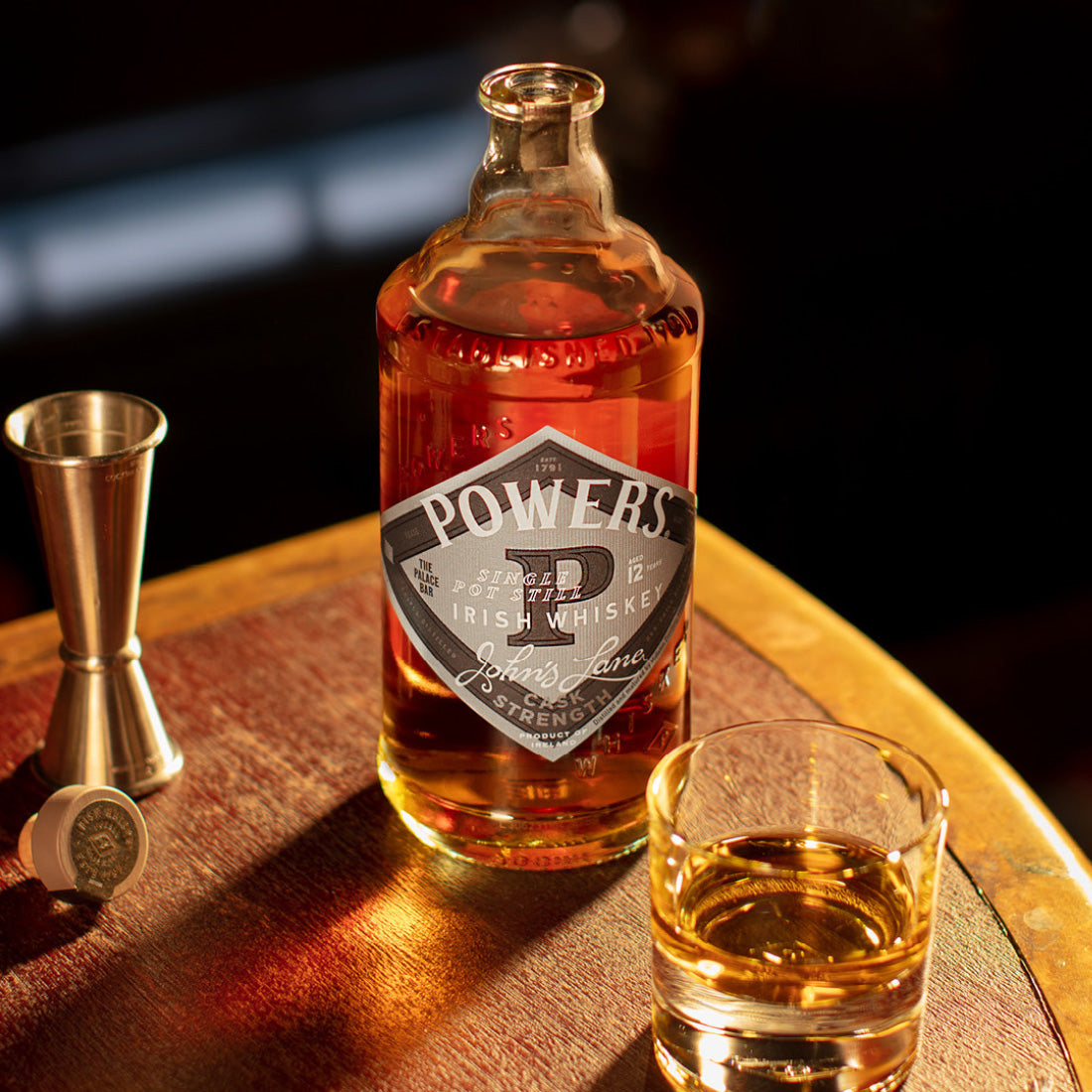 A bottle of Powers John’s Lane 12 Year Old Irish Whiskey rests on a wooden surface beside a filled glass, jigger, and cap, all illuminated by warm golden light.