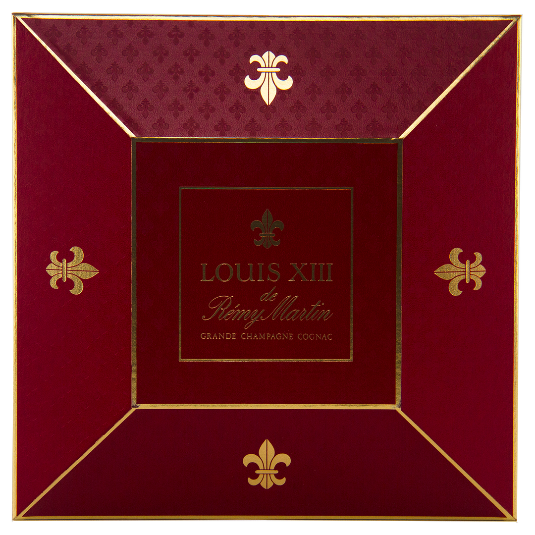 A red and gold box with "Rémy Martin Louis XIII 1980s Edition Cognac Grande Champagne 700mL" at the center, gold fleur-de-lis on each corner—crafted to house the iconic Baccarat crystal decanter of this legendary French Cognac.