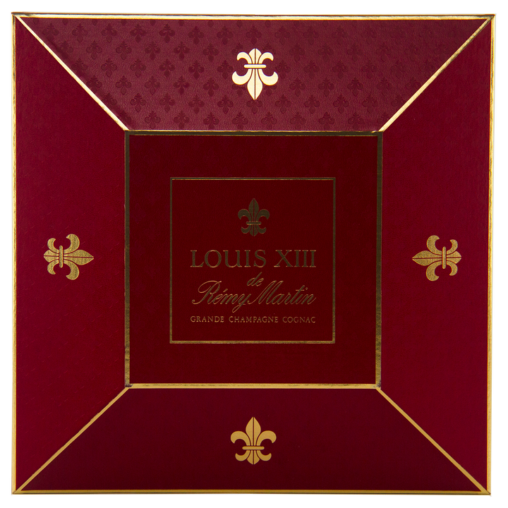 A red and gold box with "Rémy Martin Louis XIII 1980s Edition Cognac Grande Champagne 700mL" at the center, gold fleur-de-lis on each corner—crafted to house the iconic Baccarat crystal decanter of this legendary French Cognac.