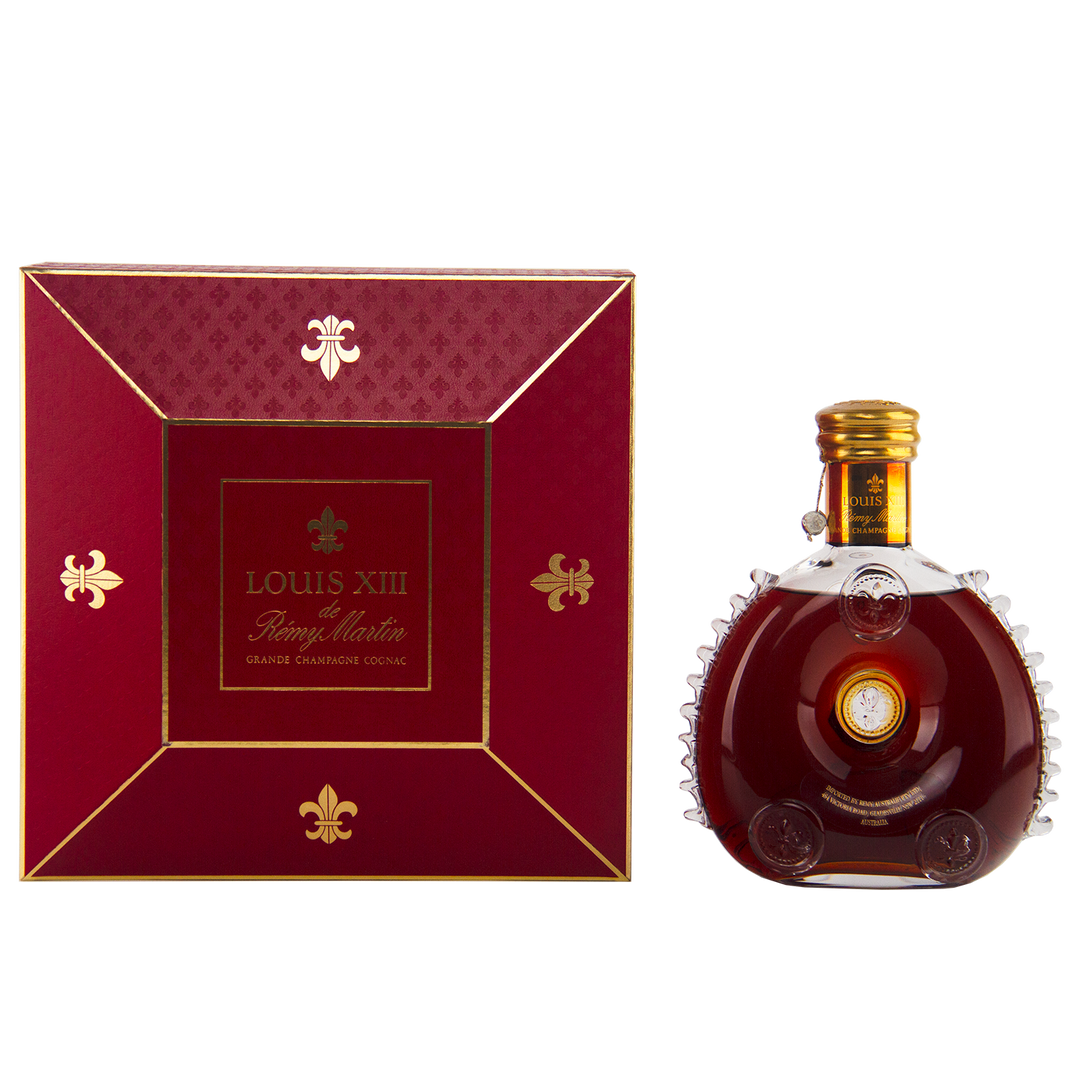 A 700mL bottle of Rémy Martin Louis XIII 1980s Edition Cognac Grande Champagne sits in a Baccarat crystal decanter beside its elegant red and gold box, both featuring the iconic branding.