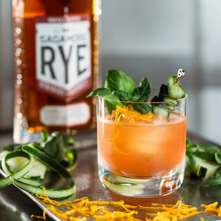 A cocktail garnished with orange zest, cucumber ribbons, and mint leaves sits on a table, with a bottle of Sagamore Spirit Cask Strength American Rye Whiskey blurred in the background.