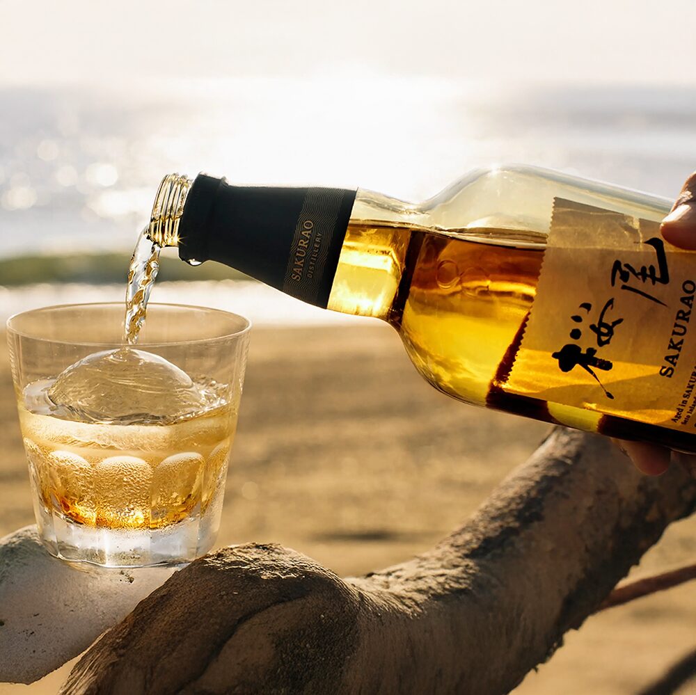 A hand pours golden Sakurao Single Malt Japanese Whisky over a large ice sphere in a glass, set on a beach as sunlight shimmers on the water—evoking the spirit of Japanese whisky by the sea.