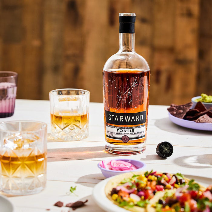 A bottle of Starward Fortis Australian Single Malt Whisky is on an outdoor table with whisky glasses, salad, pickled onions, and tortilla chips, set against a warmly sunlit wooden backdrop.