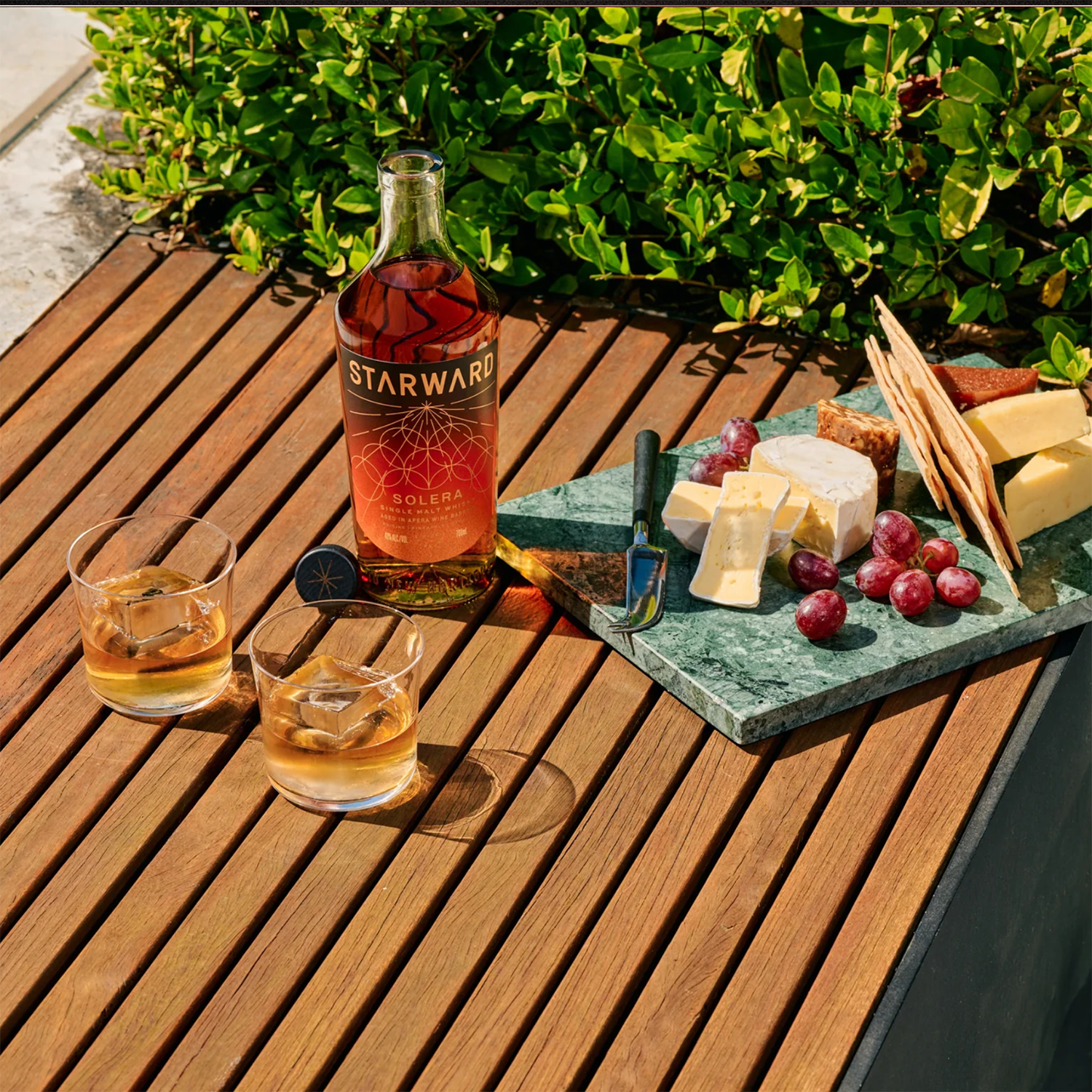 A bottle of Starward Solera Australian Single Malt Whisky, two whisky glasses with ice, and a marble platter of cheese, crackers, and grapes rest on a wooden outdoor table beside lush green bushes.