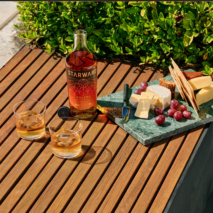 A bottle of Starward Solera Australian Single Malt Whisky, two whisky glasses with ice, and a marble platter of cheese, crackers, and grapes rest on a wooden outdoor table beside lush green bushes.