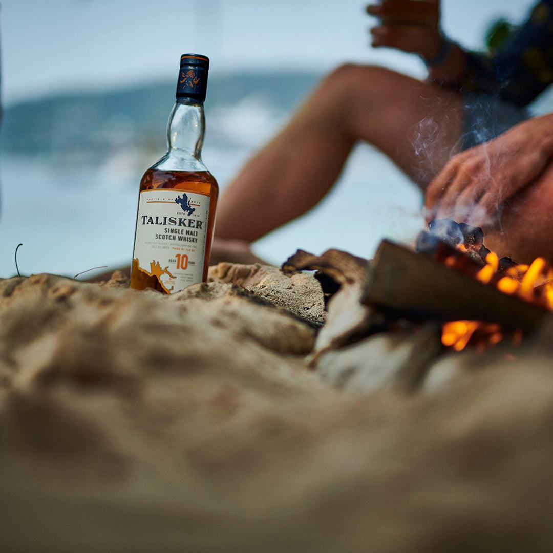A bottle of Talisker 10 Year Old Single Malt Scotch Whisky stands on sandy ground beside a campfire, with a person savoring a drink by the beach in the blurred background.