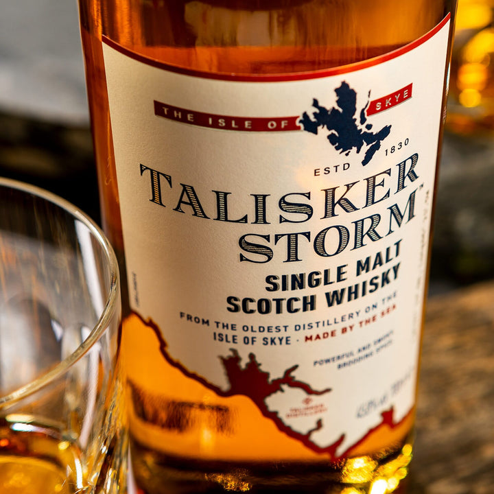 A close-up shows a bottle of Talisker Storm Single Malt Scotch Whisky with a map-detailed label, beside a glass of smoky whisky. The label emphasizes its origin from Scotland’s Isle of Skye.
