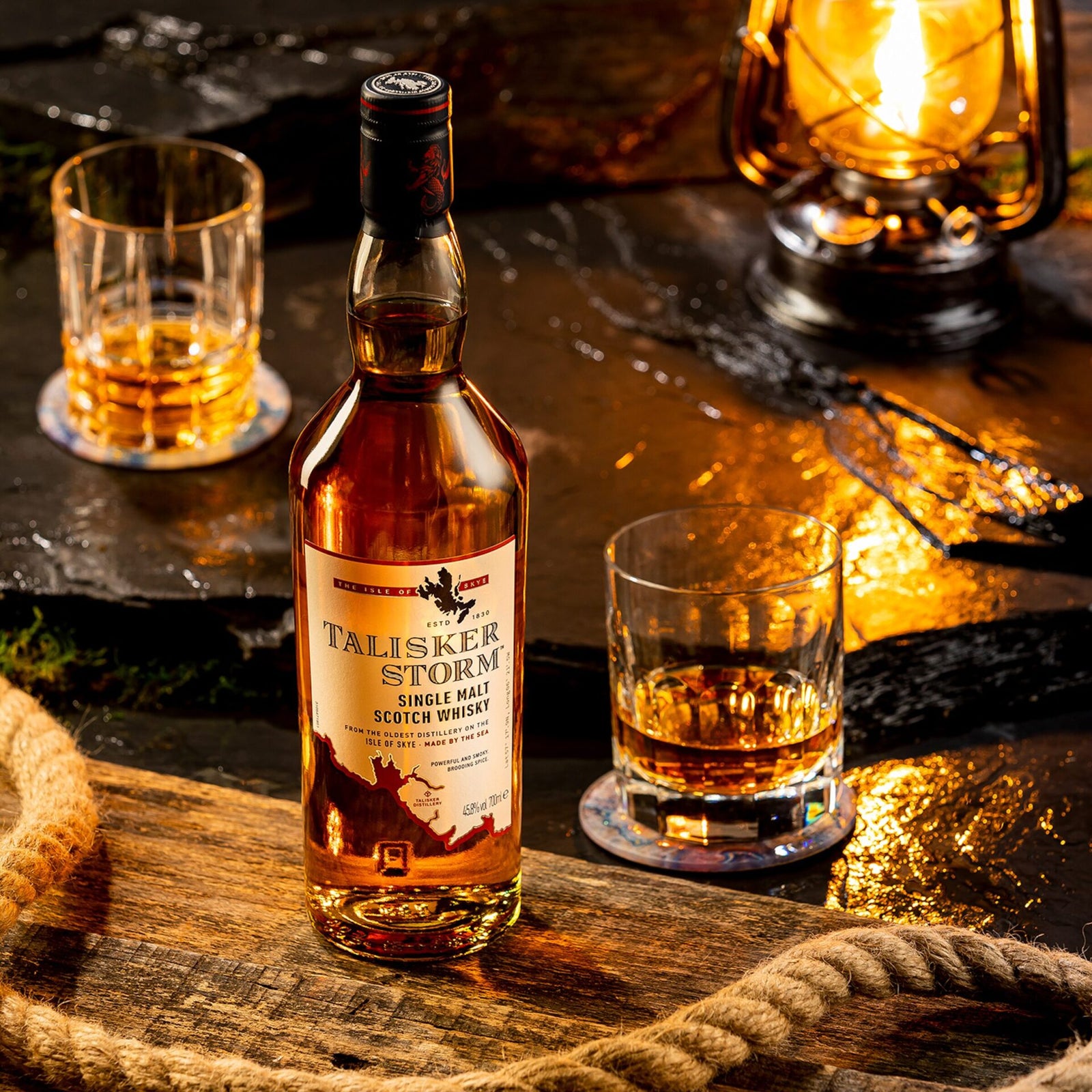 A bottle of Talisker Storm Single Malt Scotch Whisky sits on a stone surface, flanked by two glasses of amber whisky, a thick rope, and a glowing lantern in a cozy, rustic setting.