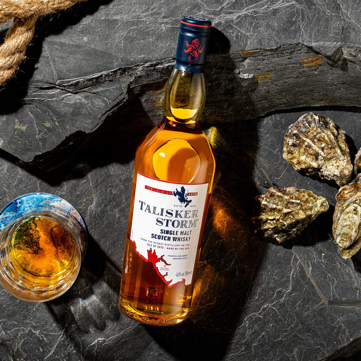 A bottle of Talisker Storm Single Malt Scotch Whisky rests on dark stone beside a glass of whisky and oyster shells, with a thick rope partly visible in the corner.