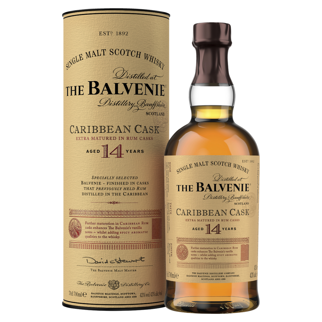 A bottle of The Balvenie Caribbean Cask 14 Year Old Single Malt Scotch Whisky stands beside its matching cylindrical box, both displaying tan labels with black and gold text.