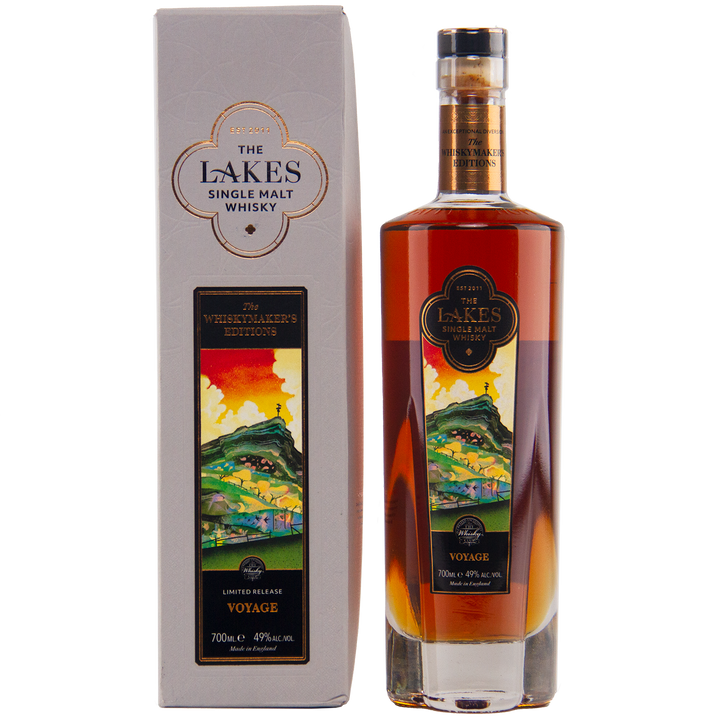 A 700ml bottle of The Lakes Whiskymaker’s Editions: Voyage Single Malt Whisky (49% ABV) stands beside its colorful scenic box. This limited release from The Lakes Distillery features hills and water on its label.