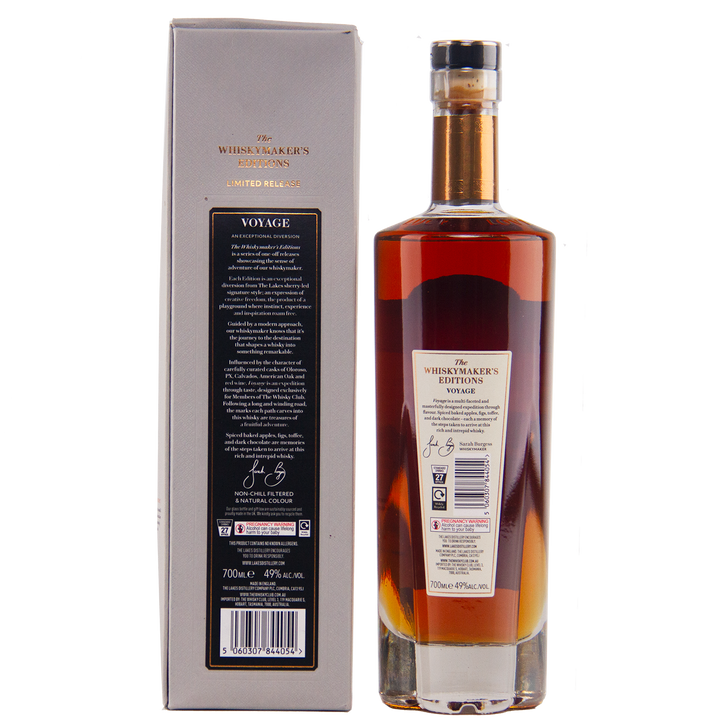 A bottle of The Lakes Whiskymaker’s Editions: Voyage Single Malt Whisky with amber liquid and a dark cap stands beside its silver-grey box, both bearing detailed labels that showcase the craftsmanship of The Lakes Distillery.