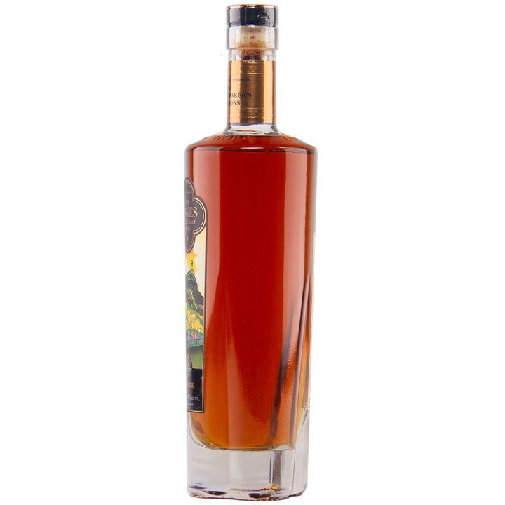 A tall, clear rectangular bottle of The Lakes Whiskymaker’s Editions: Voyage Single Malt Whisky, filled with amber liquid and sealed with a black and gold cap, features a decorative front label from The Lakes Distillery.