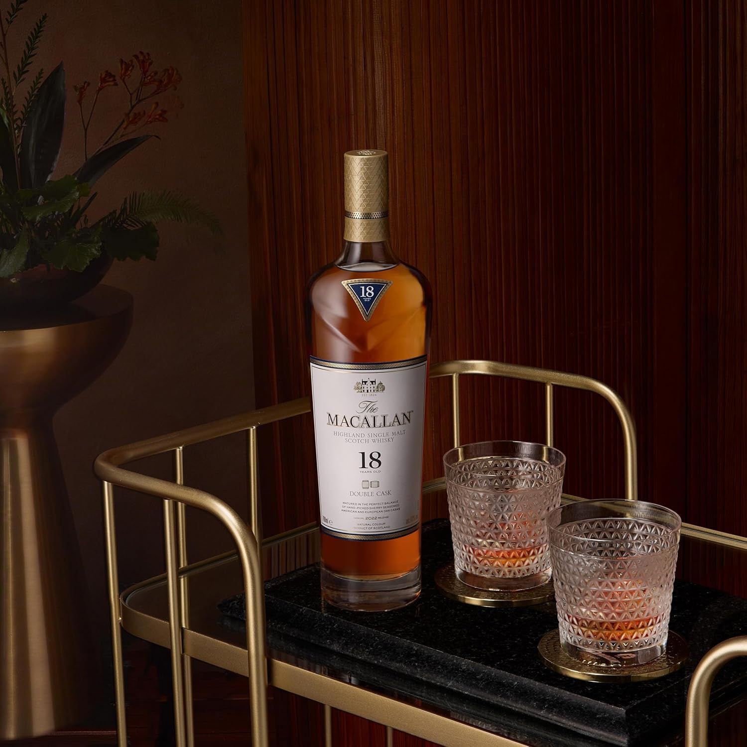 A bottle of The Macallan Double Cask 18 Year Old Single Malt Scotch Whisky rests on a gold bar cart next to two filled crystal glasses on coasters, framed by a plant and wooden wall in the background.