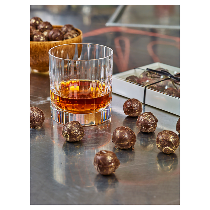 A glass of The Dalmore 12 Year Old Single Malt Scotch Whisky rests on a metal surface with chocolate truffles nearby, while more truffles in a wooden bowl and box complete the scene and complement this celebrated Scotch.