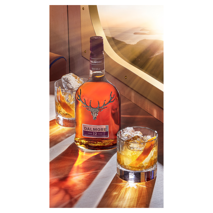A bottle of The Dalmore 12 Year Old Single Malt Scotch Whisky and two whisky glasses with ice sit on a wooden table by an airplane window, sunlight streaming in and revealing a scenic view outside.