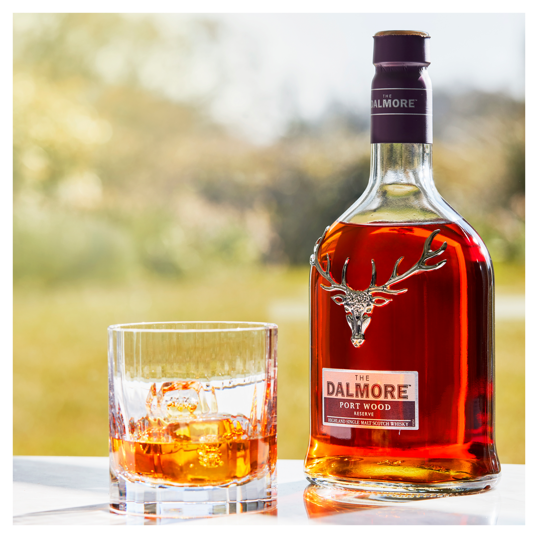 A bottle of The Dalmore Port Wood Reserve Single Malt Scotch Whisky stands next to a glass of whisky with ice on a reflective outdoor surface, set against a blurred green background, showcasing its rich port cask character.
