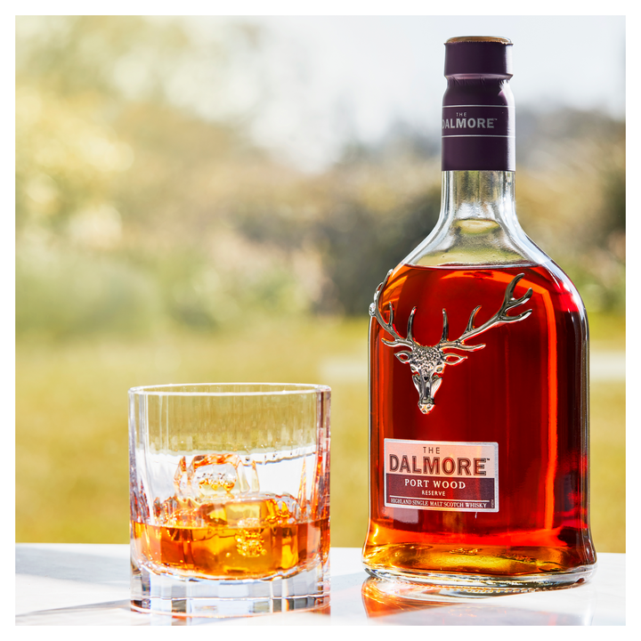 A bottle of The Dalmore Port Wood Reserve Single Malt Scotch Whisky stands next to a glass of whisky with ice on a reflective outdoor surface, set against a blurred green background, showcasing its rich port cask character.