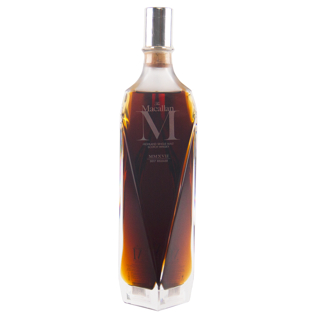 A tall, faceted clear glass bottle of The Macallan M (2017 First Release) Single Malt Scotch Whisky with dark amber liquid and a silver cap—an elegant showcase of sherry oak maturation, set against a plain white background.