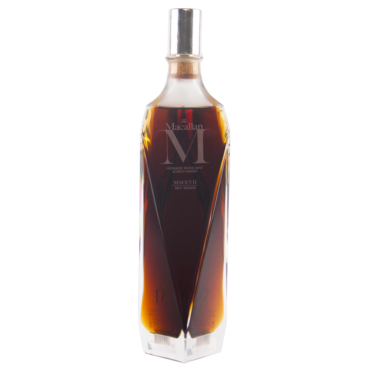 A tall, faceted clear glass bottle of The Macallan M (2017 First Release) Single Malt Scotch Whisky with dark amber liquid and a silver cap—an elegant showcase of sherry oak maturation, set against a plain white background.