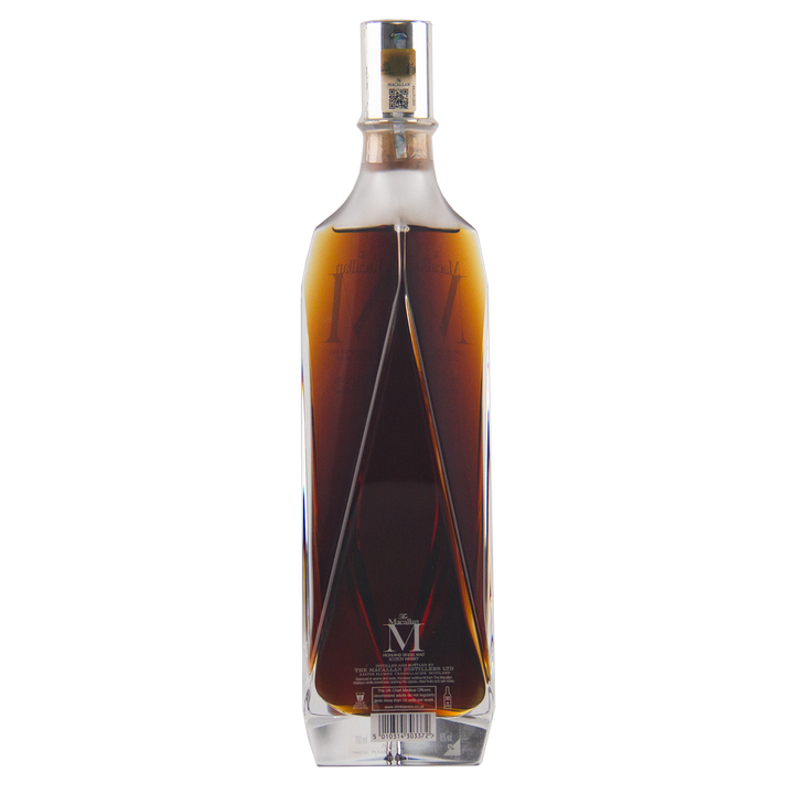 A bottle of The Macallan M (2017 First Release) Single Malt Scotch Whisky in a geometric clear crystal decanter with dark amber liquid, displayed against a white background and highlighting its rich sherry oak maturation.