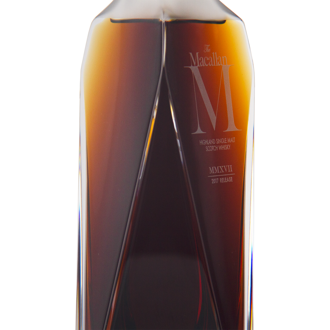 A close-up of The Macallan M (2017 First Release) Single Malt Scotch Whisky highlights its dark amber liquid in a clear, angular decanter with engraved text, emphasizing its sherry oak maturation.
