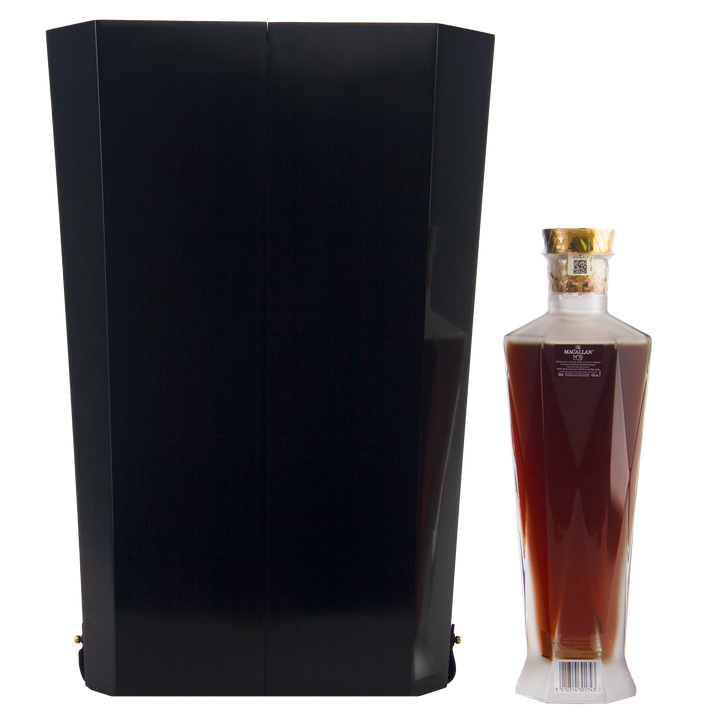 A tall black box stands beside an elegant, angular bottle of The Macallan No 6 Single Malt Scotch Whisky, filled with dark amber liquid and topped with a gold cap, both set against a plain white background.