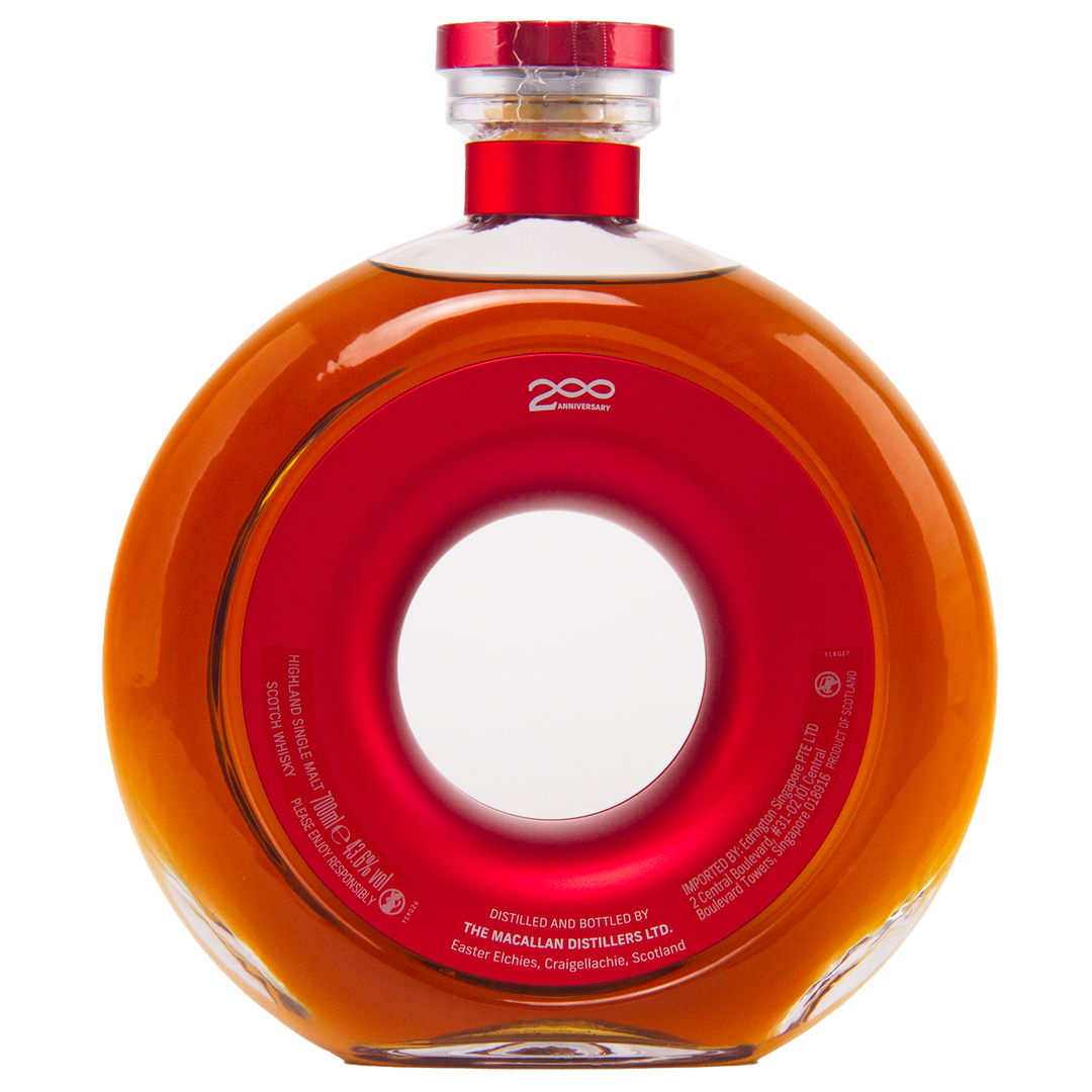 A round, clear glass bottle of The Macallan Time Space Mastery Single Malt Scotch Whisky with a central hole, filled with amber liquid and topped with a red and gold cap. The label honors The Macallan 200th Anniversary—a collectible from Scotland.
