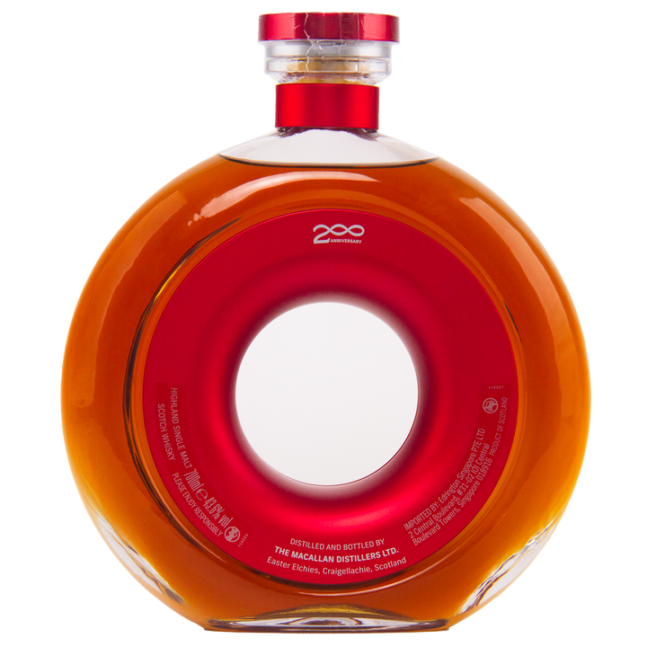 A round, clear glass bottle of The Macallan Time Space Mastery Single Malt Scotch Whisky with a central hole, filled with amber liquid and topped with a red and gold cap. The label honors The Macallan 200th Anniversary—a collectible from Scotland.