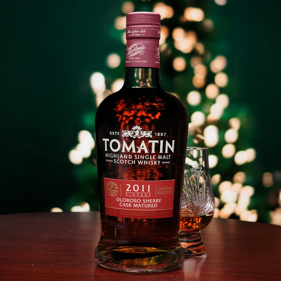 A bottle of Tomatin Highland Single Malt Scotch Whisky with a 2011 Collection label, standing on a wooden table next to a small crystal whisky glass. The background features a dark green wall with warm, out-of-focus bokeh lights.