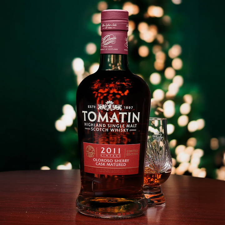 A bottle of Tomatin Highland Single Malt Scotch Whisky with a 2011 Collection label, standing on a wooden table next to a small crystal whisky glass. The background features a dark green wall with warm, out-of-focus bokeh lights.